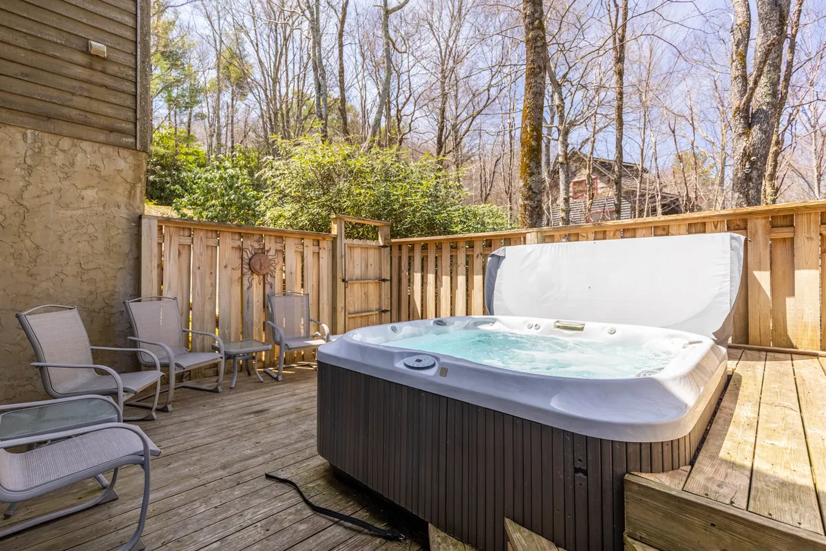A spacious wooden deck nestled among tall trees features a bubbling hot tub, lounge chairs, and serene views of the surrounding forest. The inviting setting merges seamlessly with nature, offering a tranquil space to relax after a day of outdoor adventure.