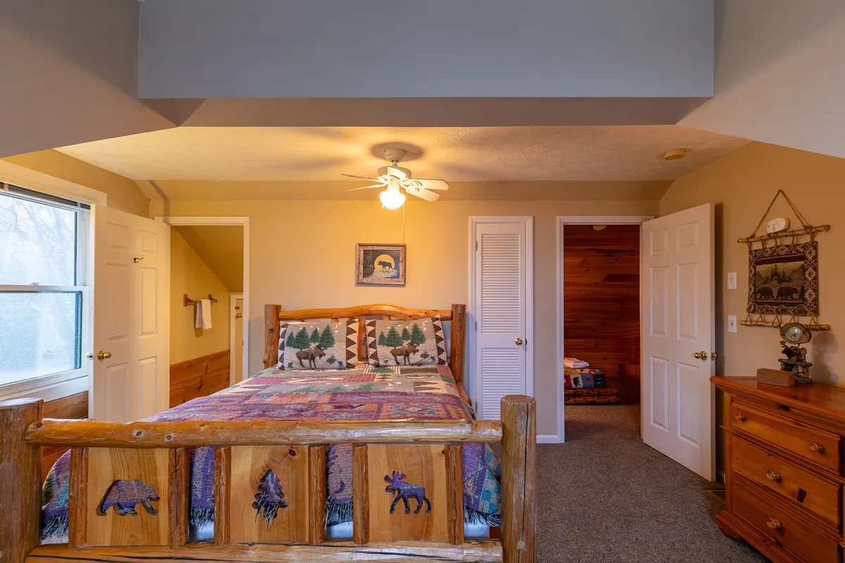 A cozy bedroom with a log bed featuring decorative bedding, wooden furniture, and framed artwork. The room exudes a welcoming, restful vibe with natural light streaming in. The hand-crafted log details are visible, matching the mountain artistry described for the queen bedroom.