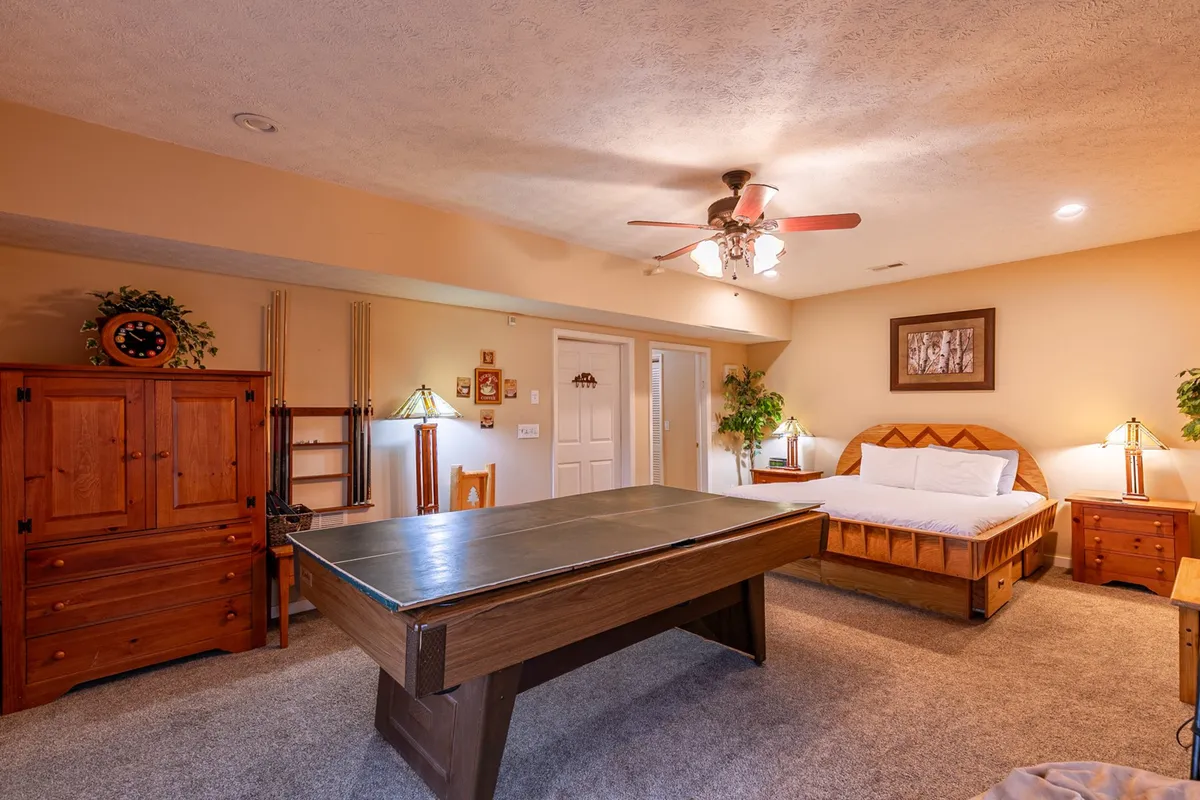 A downstairs recreation room featuring a pool table as the focal point, complemented by a sofa bed, wooden furniture, and warm lighting from cozy lamps. The space feels casual and inviting, ready for games, relaxation, or group entertainment.