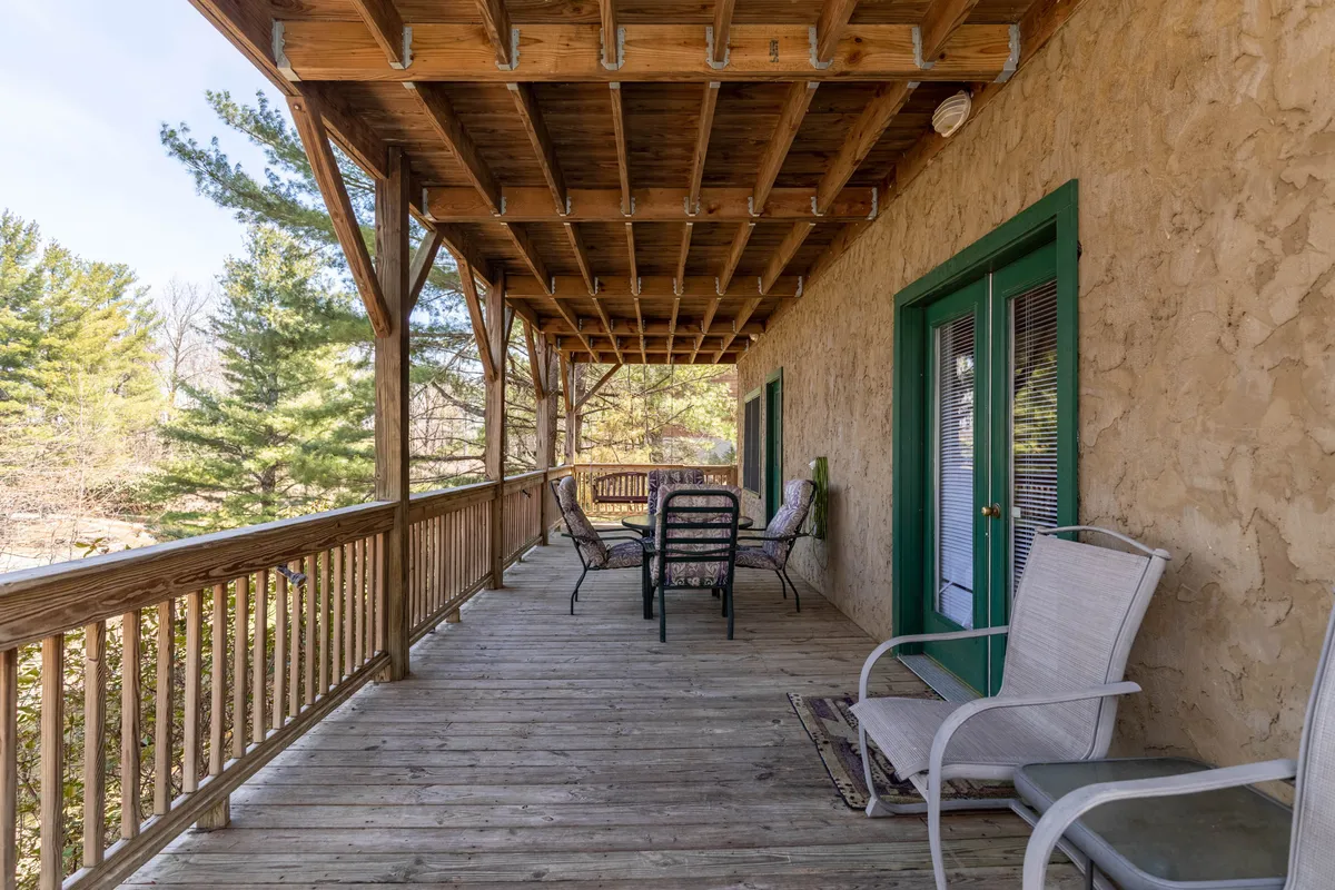 A spacious wooden deck with green doors, surrounded by trees and featuring comfortable outdoor seating. The atmosphere is tranquil, inviting guests to relax, socialize, or enjoy a quiet morning coffee while soaking in the natural environment.