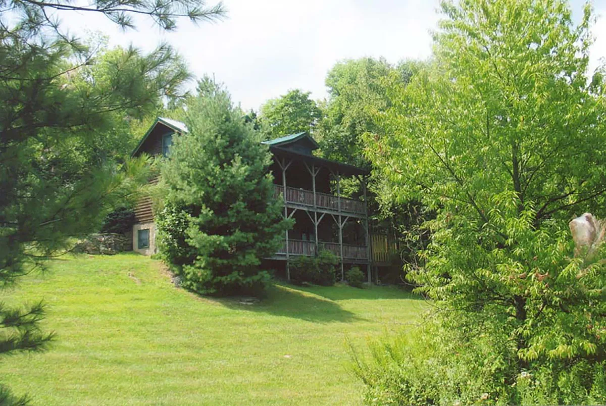 The image captures a charming rustic cabin nestled in lush greenery, featuring a spacious wooden balcony overlooking a sloping lawn. The serene setting is perfect for relaxing on the porch, enjoying breathtaking sunsets, and gathering with loved ones for outdoor activities, all set against the majestic backdrop of the Blue Ridge Mountains.