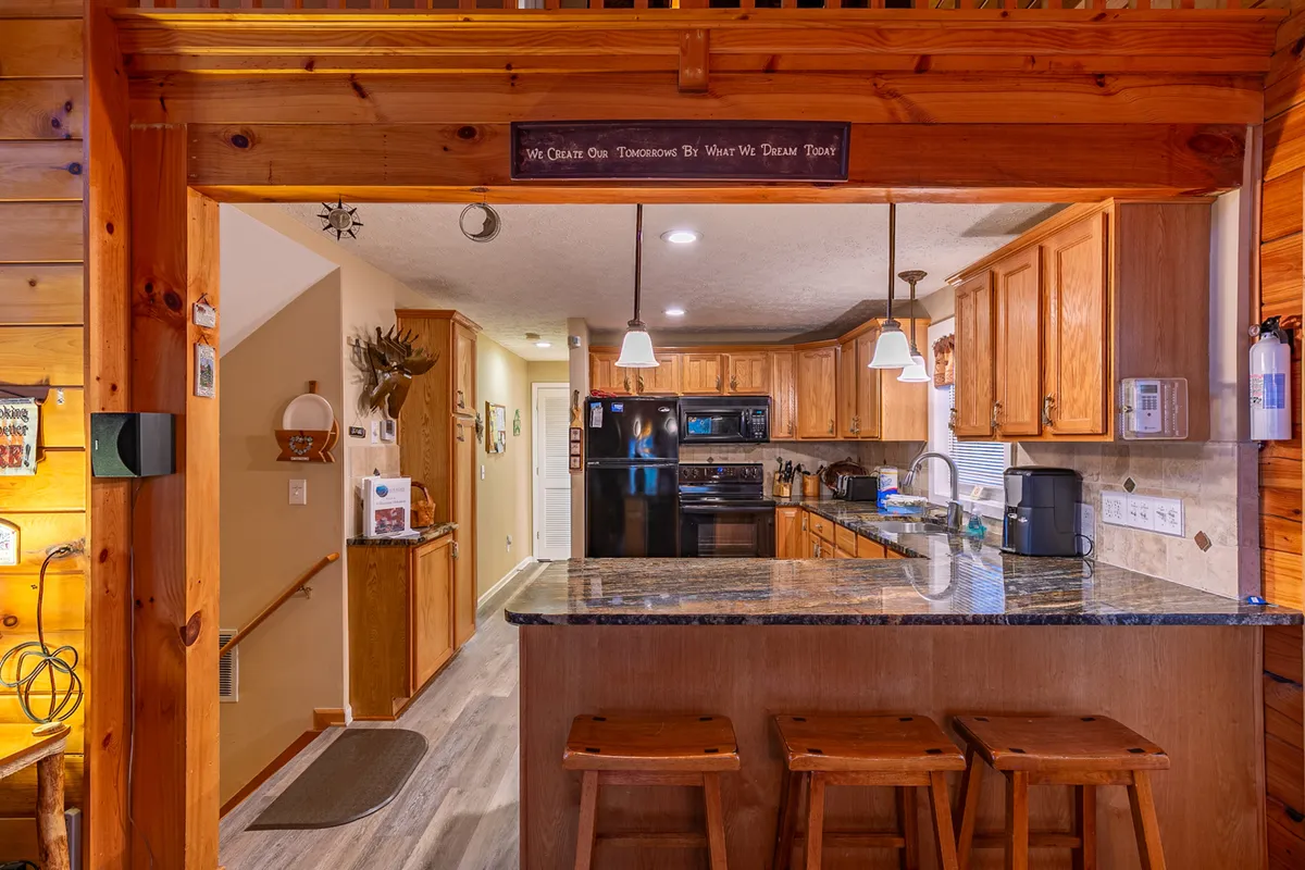 A kitchen scene featuring a granite countertop with stools, wooden cabinetry, and pendant lighting. The arrangement suggests a space well-suited for entertaining and group cooking, with enough room for people to gather, cook, and share meals.