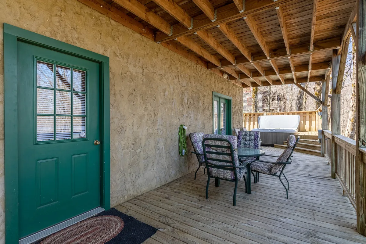 This image captures a spacious exterior deck area featuring a dining table with chairs in the foreground and a hot tub set in the background. The green door leads back inside, while the scene is framed by mountain views and sunset lighting, suggesting both calm and social possibilities. The setting naturally invites outdoor dining, socializing, or relaxing in the hot tub—all under a covered deck.