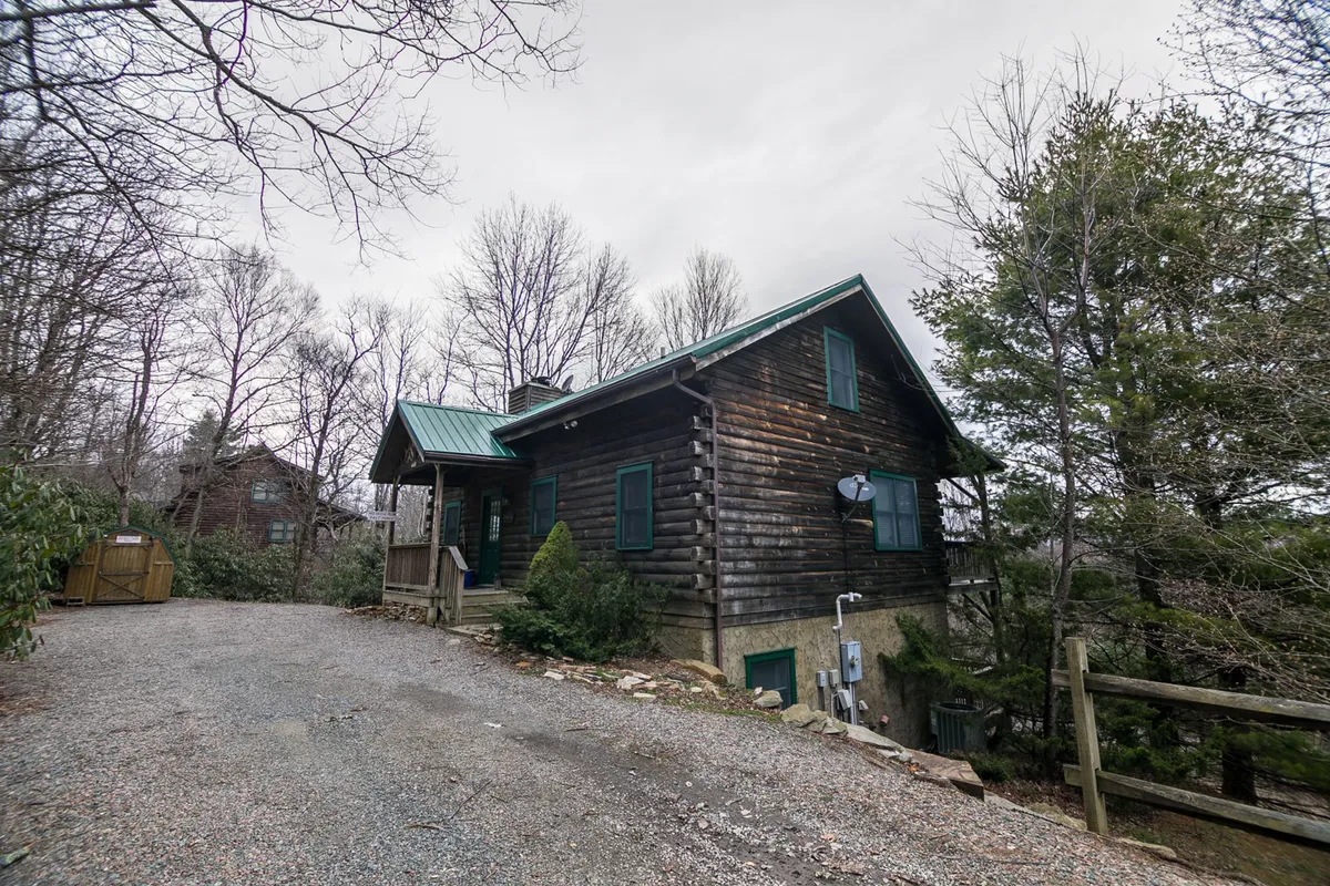 An inviting exterior view of the log cabin, complete with a green roof, surrounded by mature trees and a gravel driveway. The rustic setting evokes a sense of seclusion and tranquility in the Blue Ridge mountains.