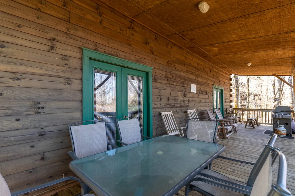 An outdoor seating area featuring a glass table, green doors, and wooden walls. The space is set up for outdoor dining or relaxing with a morning coffee, connecting the interior comfort with the scenic outdoors.