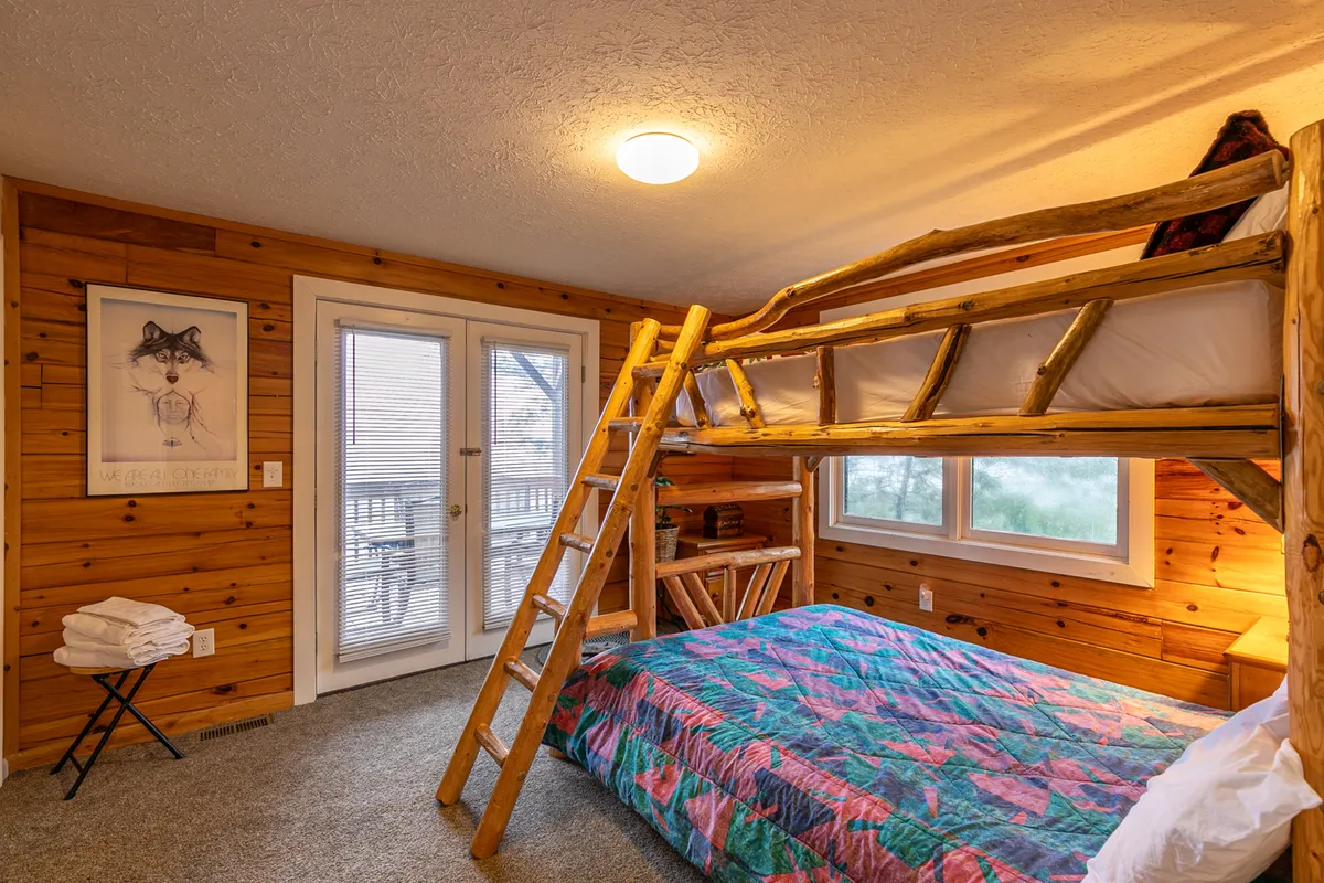 A playful bunk bed setup with a colorful quilt, wooden accents, and expansive windows that let in sunlight. The mood is cheerful and relaxed, suitable for families or groups, and the setting fits the description of a handmade log bunk bed room.
