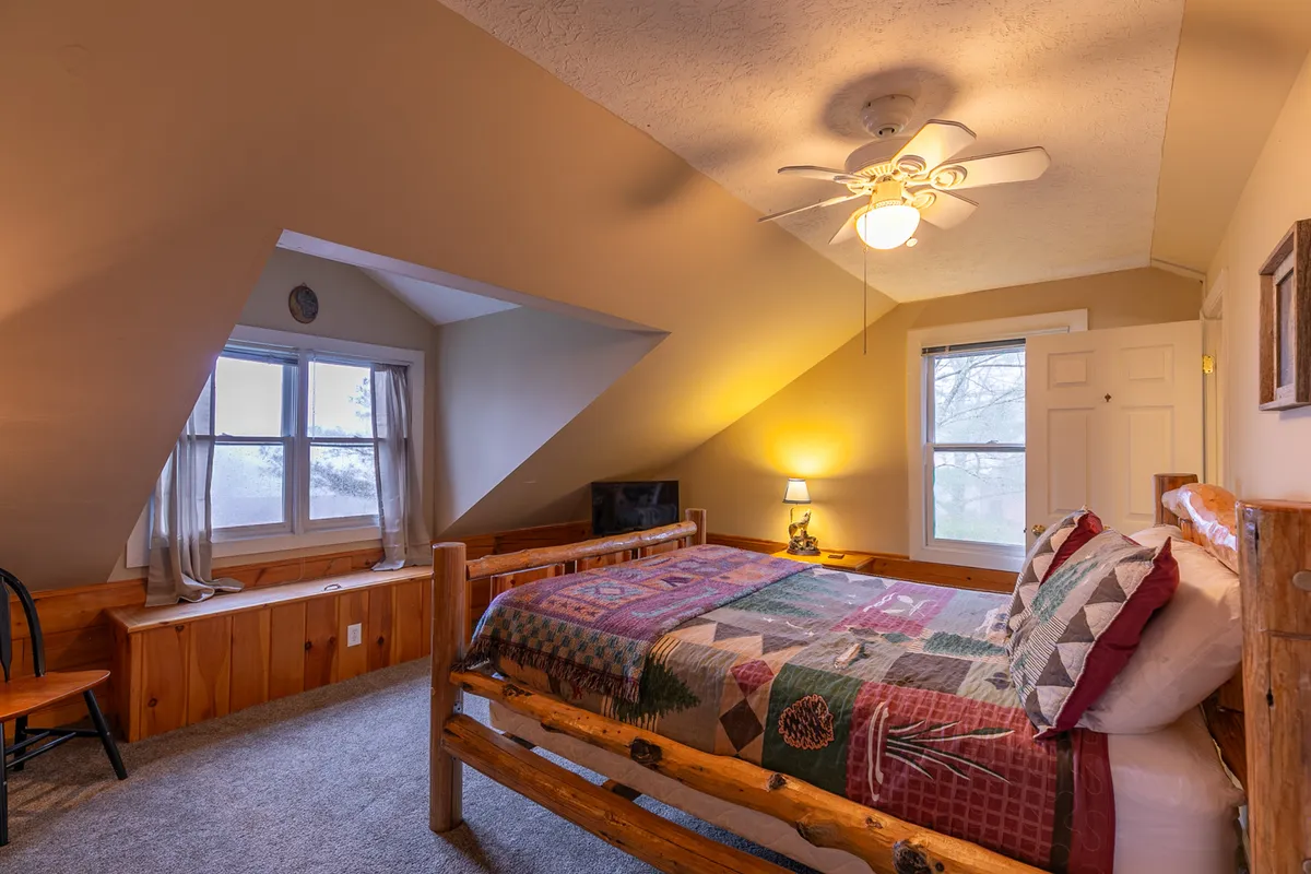 A warmly lit bedroom with a wooden bed frame, patchwork quilt, window with natural light, and cozy furnishings including a chair and nightstand. The room feels inviting and perfect for a restful night or peaceful morning, suitable for a loft or additional sleeping space.
