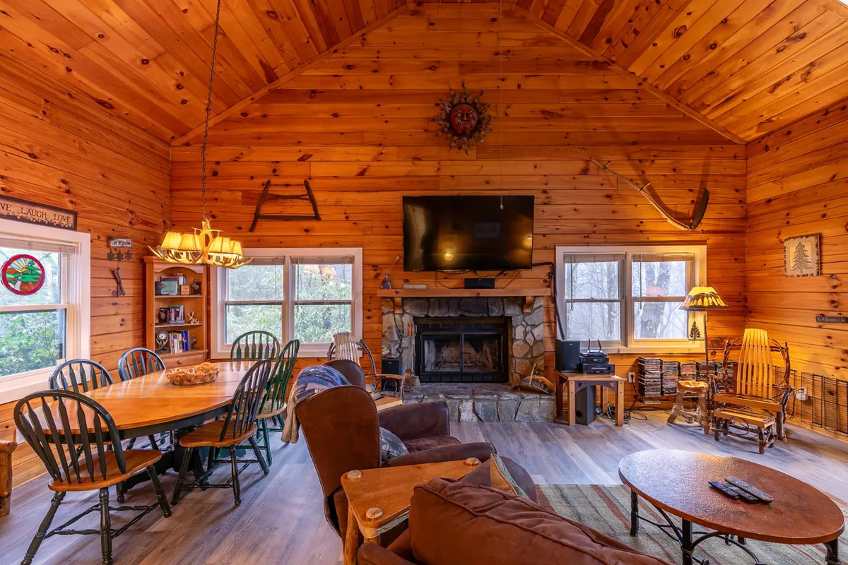 This interior scene captures an open area where the fireplace, dining table, exposed wooden beams, and a television are all visible. The space is set up for group gatherings, with enough room for multiple people to sit, play games, or watch movies together. The ambiance is warm and welcoming, thanks to the wood finishes and inviting lighting.